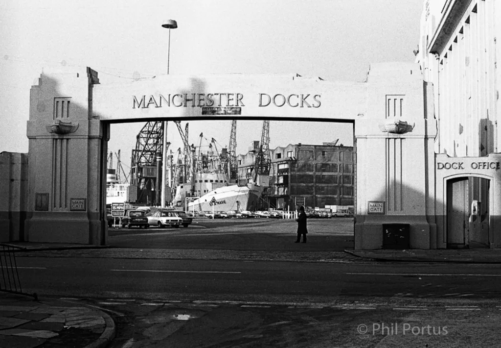 A black and white view through an arched gateway reveals a large industrial area with cranes and vehicles.