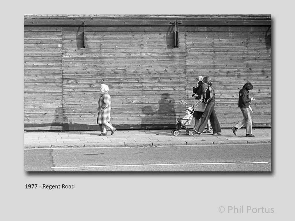 Three figures walk along a sidewalk next to a wooden wall in black and white.