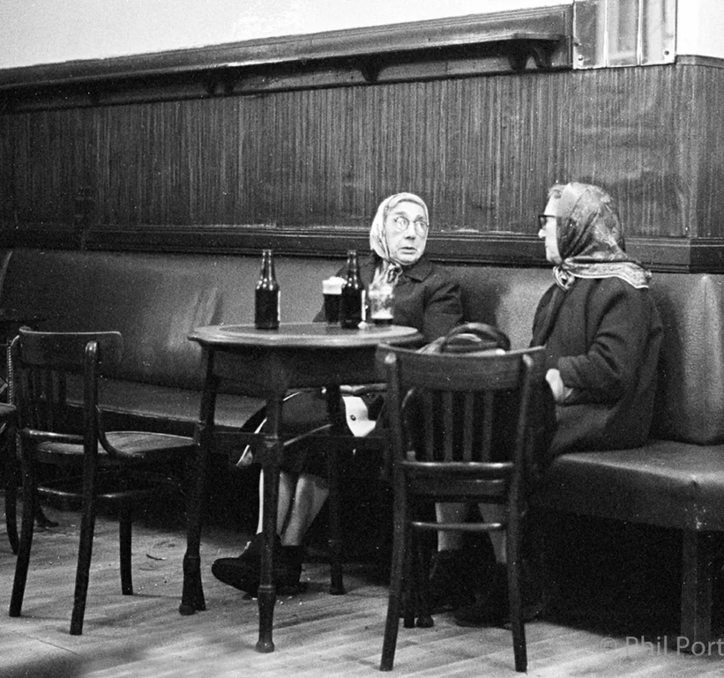 Two women sit at a small table in a dimly lit booth with bottles and glasses.