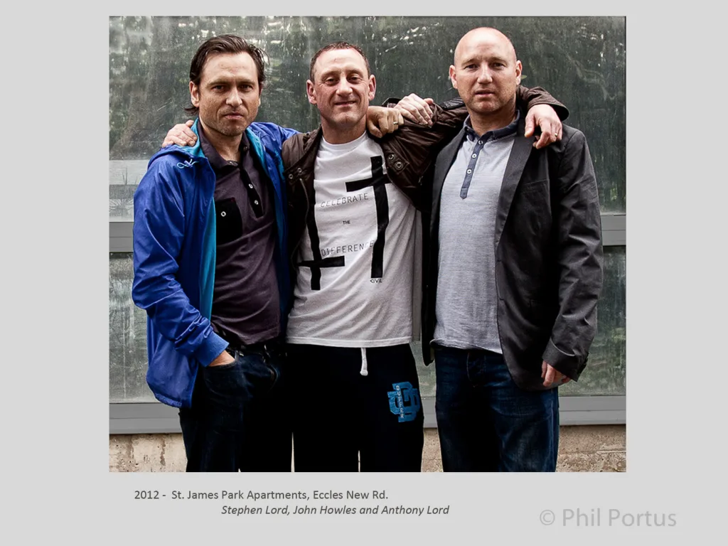Three men stand close together outdoors, arms around each other, in casual clothing.