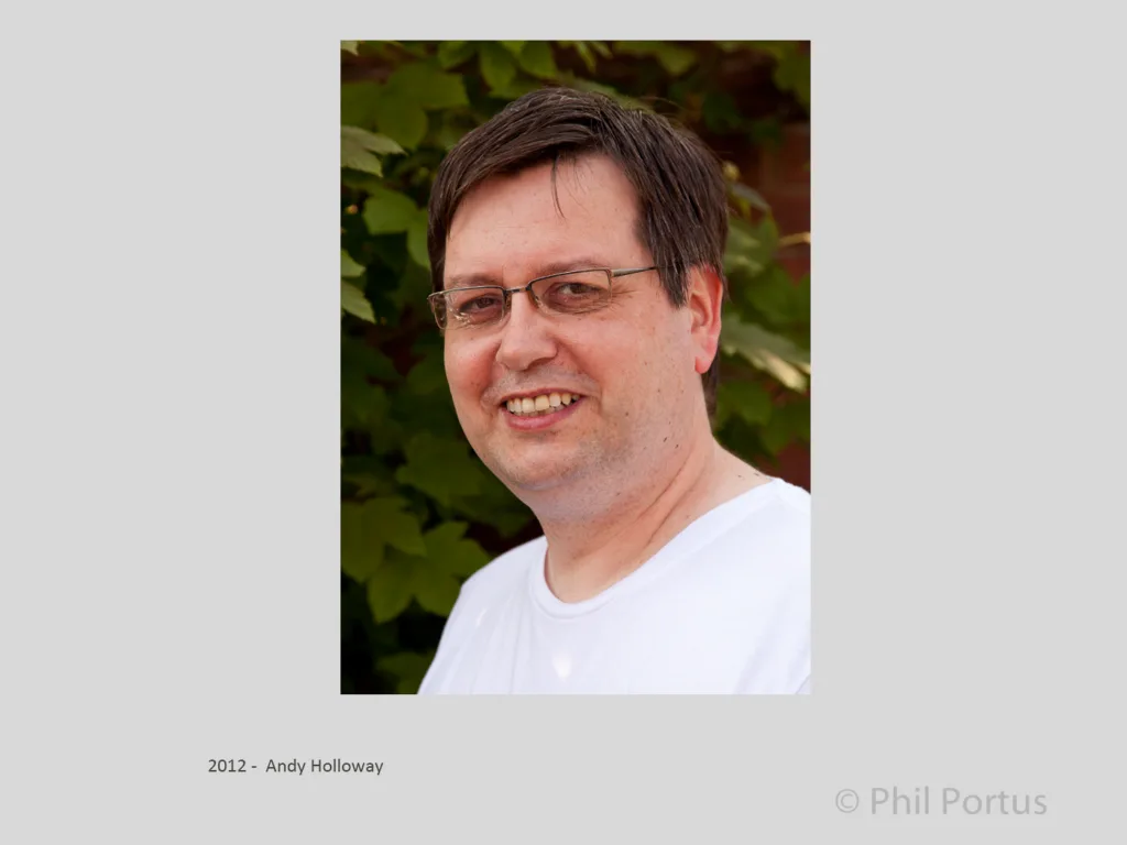 A man with glasses smiles at the camera while standing in front of green foliage.