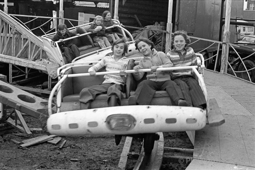 Three young people ride a spinning amusement park ride while others watch in the background.