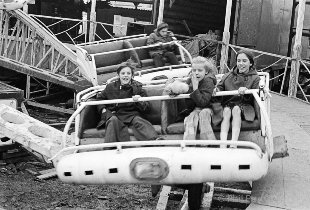 Three children sit in a vintage amusement ride car amidst other damaged rides in black and white.