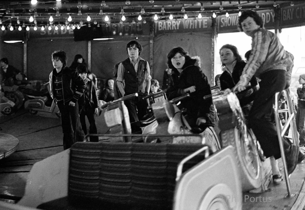 A black and white scene shows a group of people on an indoor amusement ride with string lights overhead.