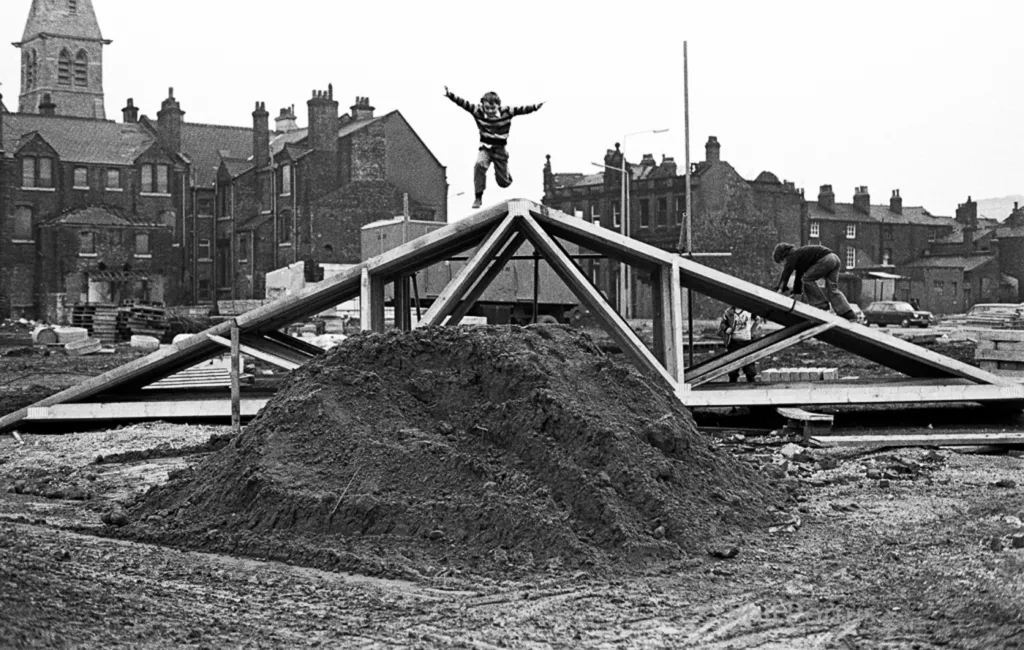 A black and white image shows a triangular wooden structure on a mound of dirt with figures nearby.