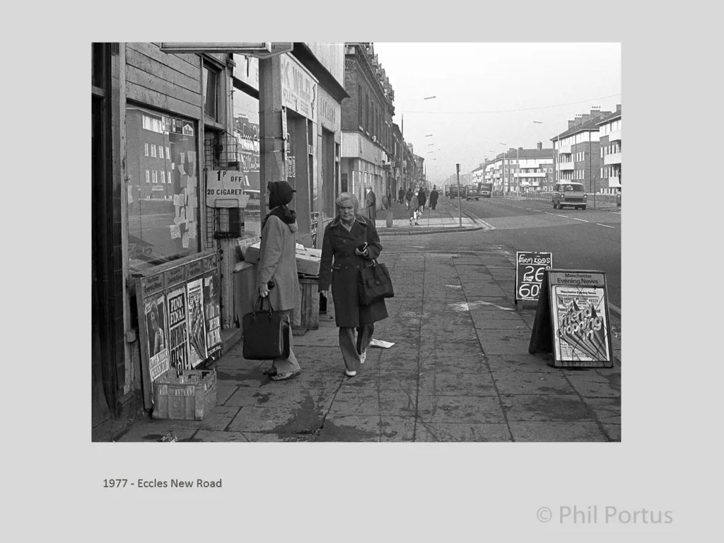 Two people stand near a shopfront with posters; another person walks along a sidewalk on a street.