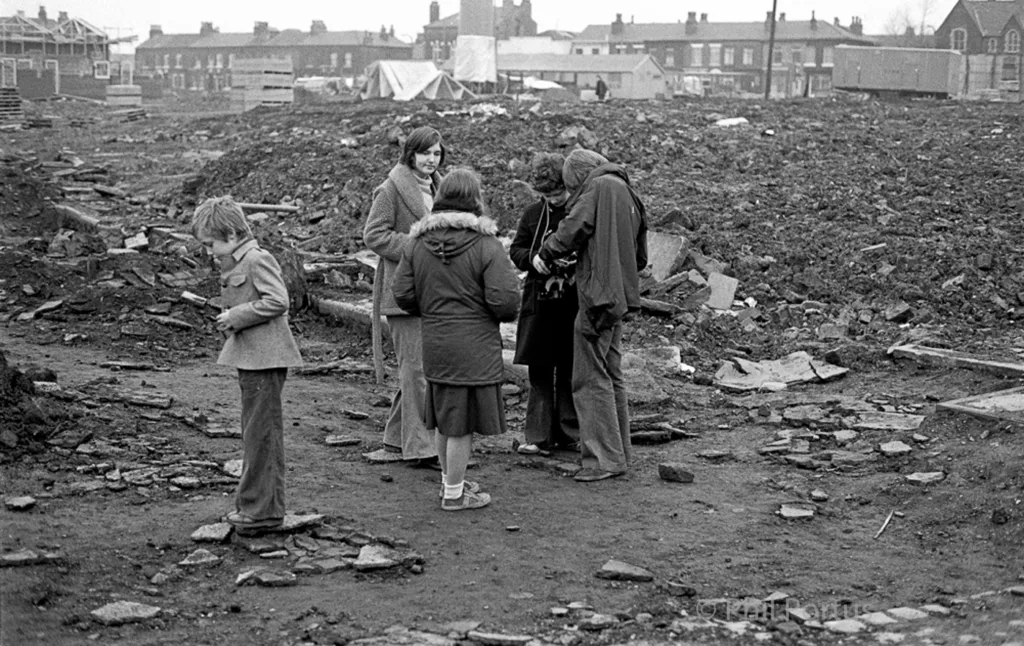 Black and white image shows four children and a woman standing in rubble surrounded by demolished buildings.