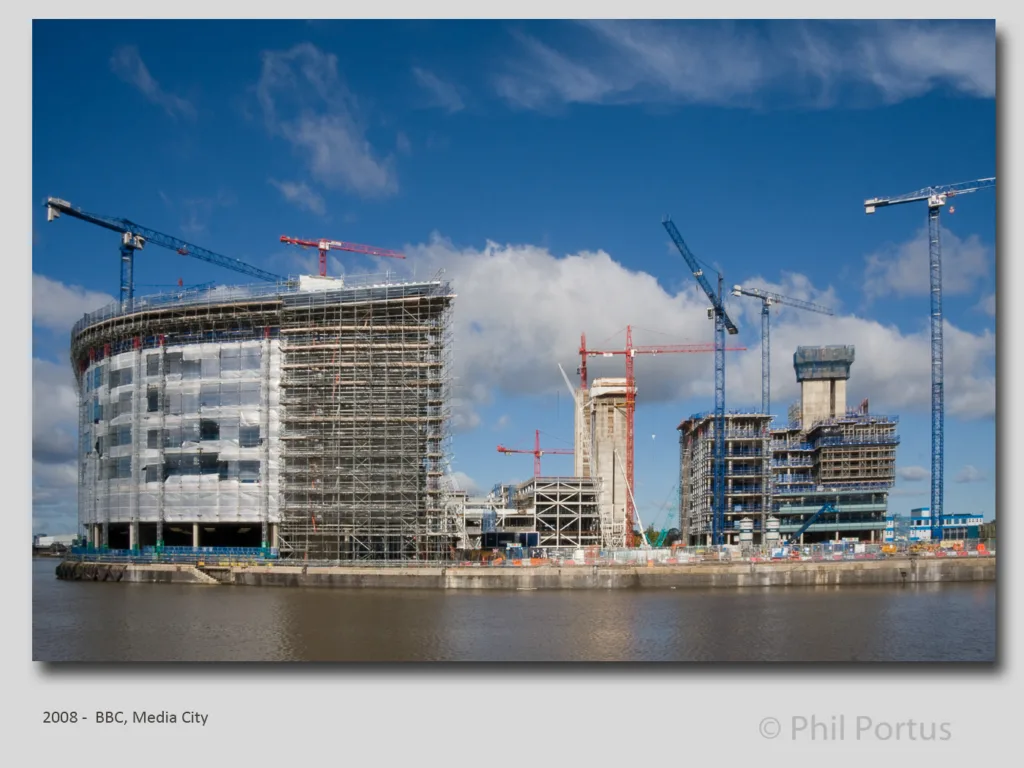 Several tall construction cranes surround partially completed buildings along a body of water under a partly cloudy sky.