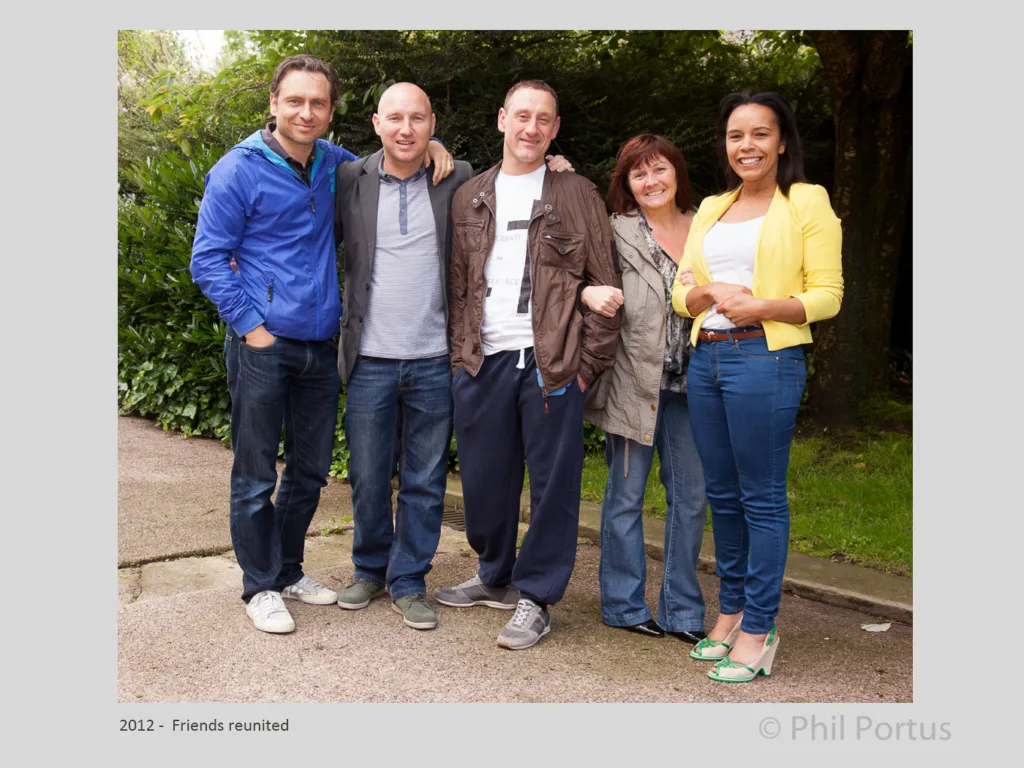 Five adults stand together outdoors on a paved surface with greenery in the background.