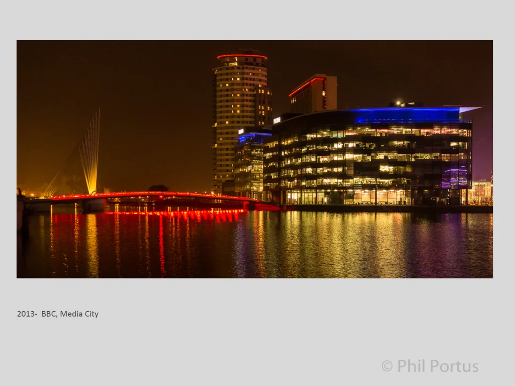Illuminated buildings reflect on dark water beneath a brightly lit red bridge at night.
