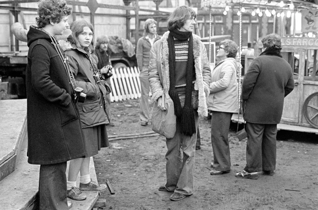A black and white scene shows a group of people gathered in an outdoor setting with a fairground structure visible.