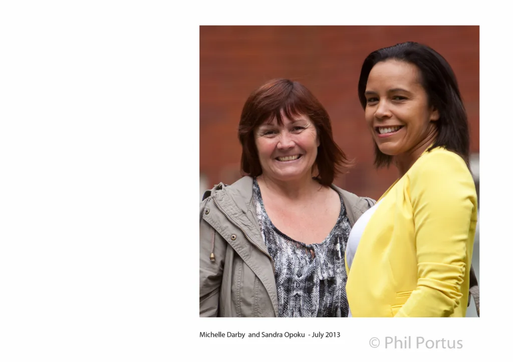 Two smiling women stand close together outdoors, one in a yellow jacket and the other in a patterned top.
