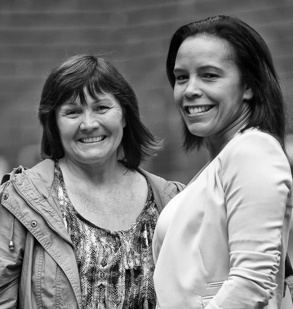 Two smiling women stand close together, one arm around the other's shoulder in a black and white portrait.