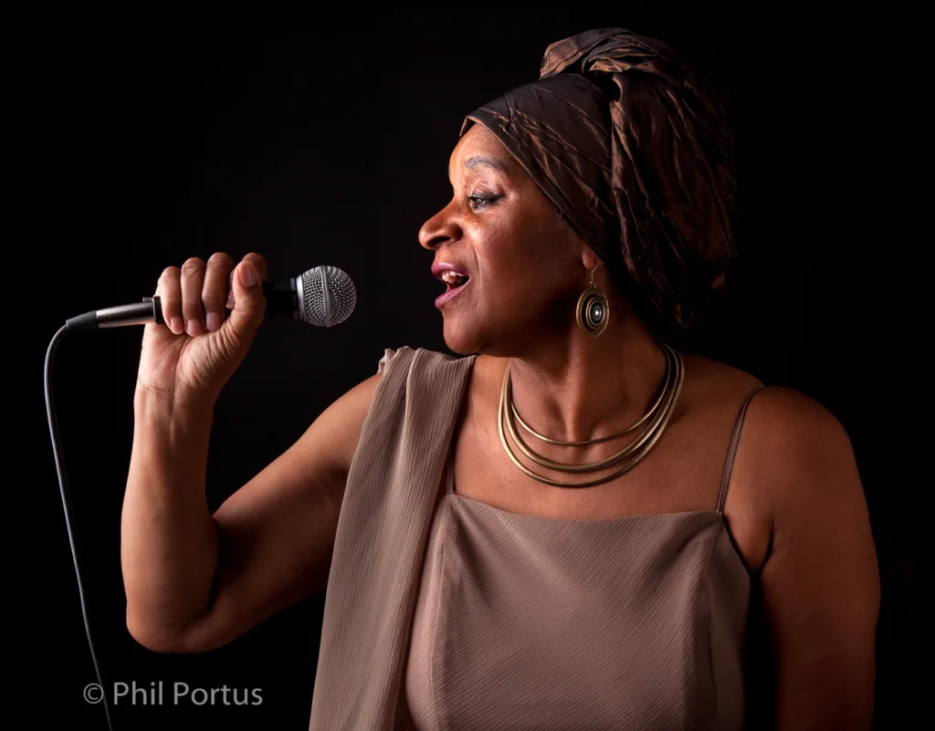 A woman with a headwrap sings into a microphone against a dark background.