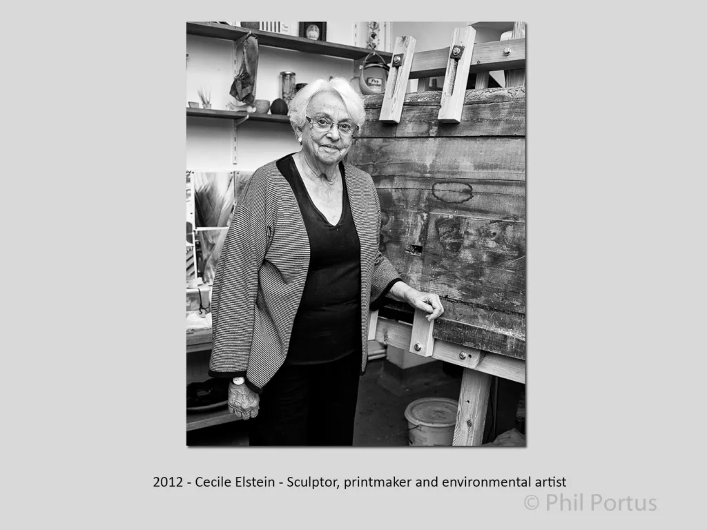 A senior woman stands near a large wooden printing press in a black and white portrait.