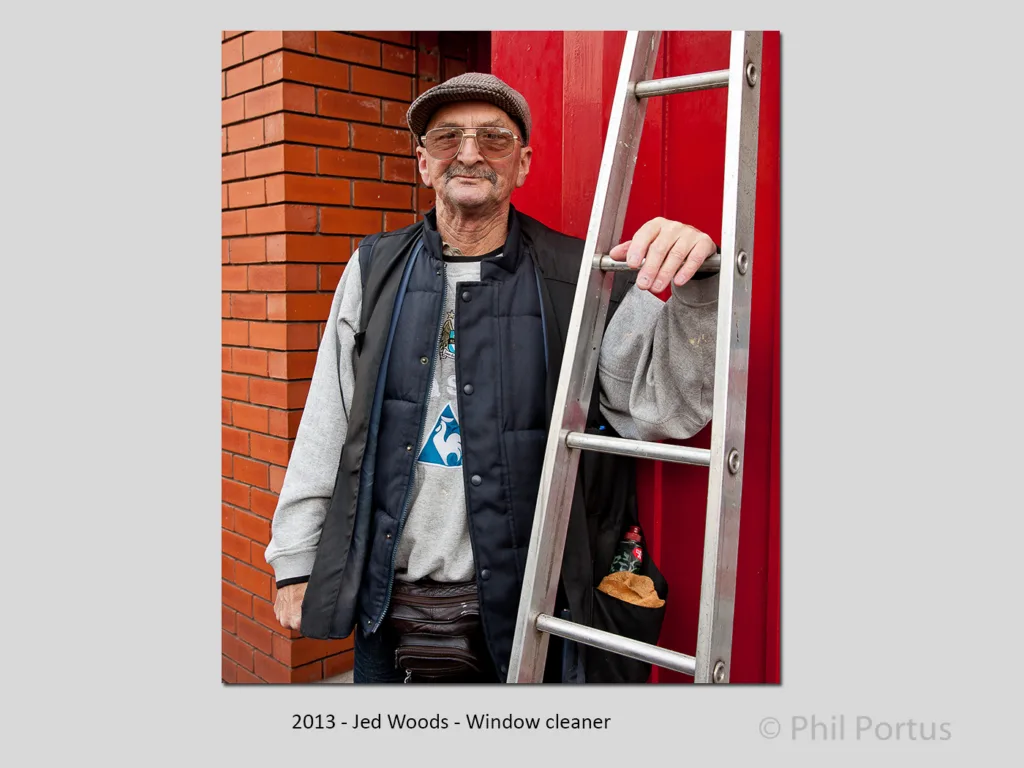 An older man wearing a cap, sweater, vest, and leather pants leans against a red door with a ladder.