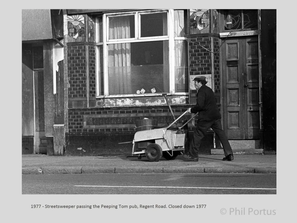 A person pushes a wheeled street cleaning machine past a brick building with boarded windows.