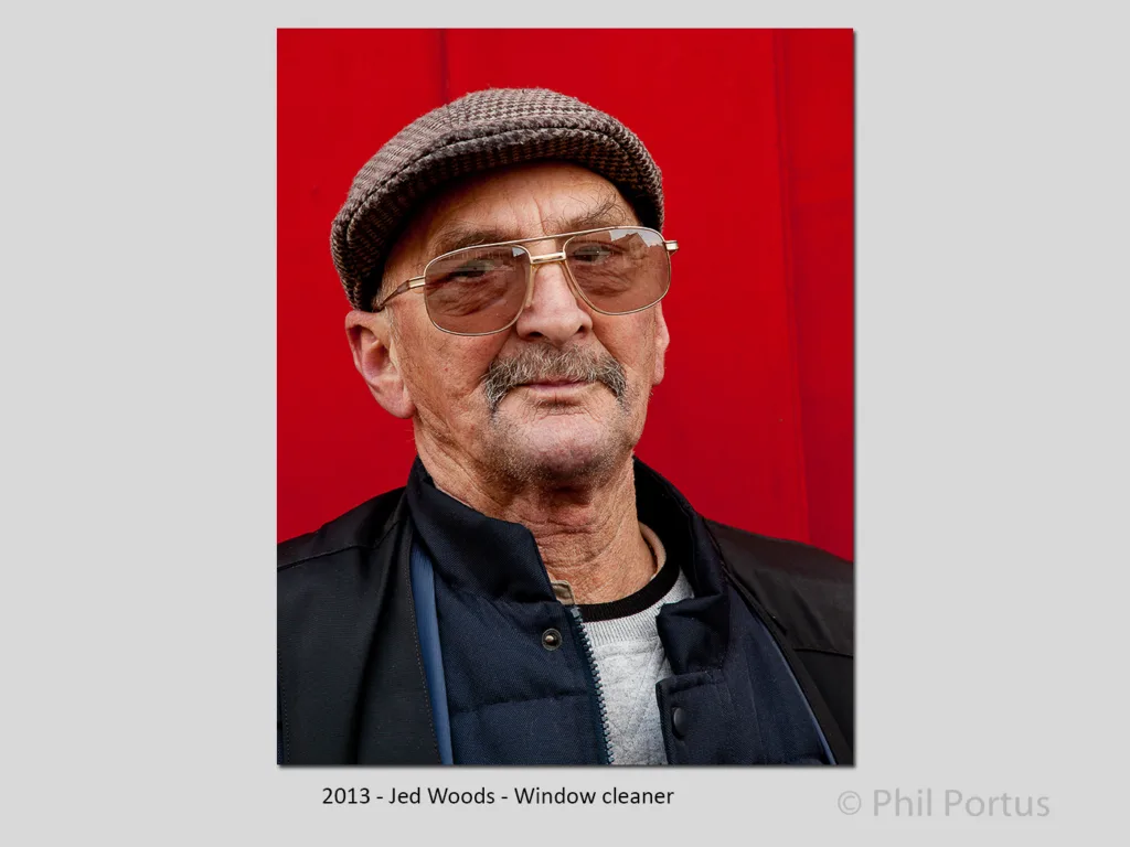 An older man wearing glasses, a hat, and a jacket poses against a bright red background.