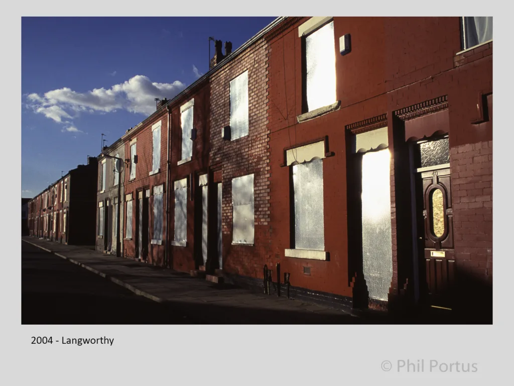 A row of brick buildings with boarded windows faces a paved walkway under a partly cloudy sky.