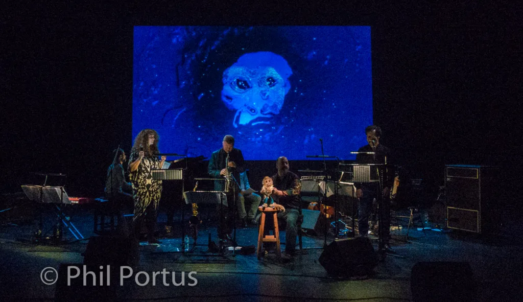 A stage is lit with blue light, featuring a group of musicians and a large projected image.