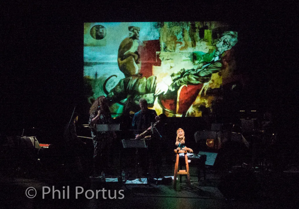 A stage performance features musicians seated before music stands with a projected image behind them.