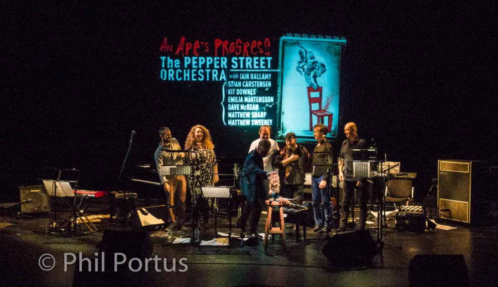 A group of musicians stands on a stage in front of a dark backdrop with text and an illustration.
