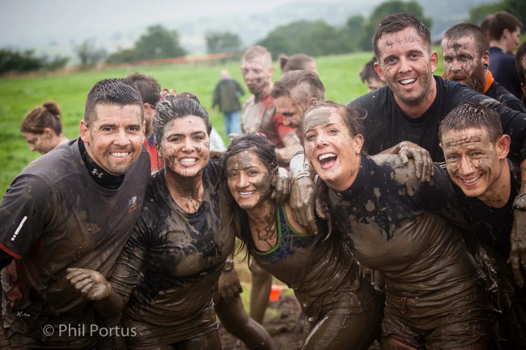 A group of people covered in mud smile for the camera amidst a grassy field.