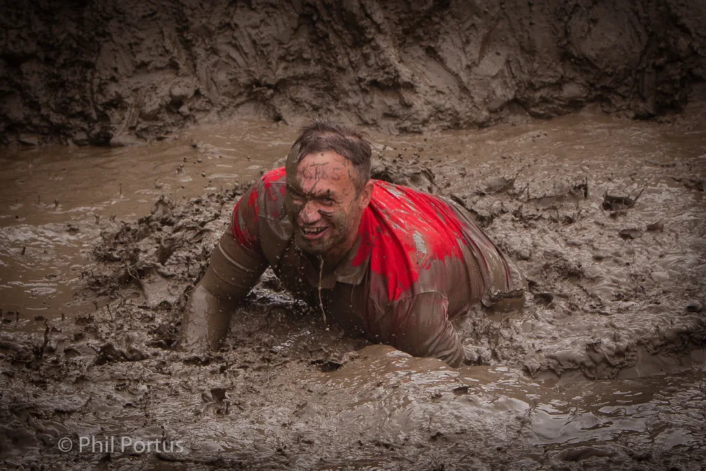 A person struggles through thick mud, covered in dirt and splashes, with a red shirt.