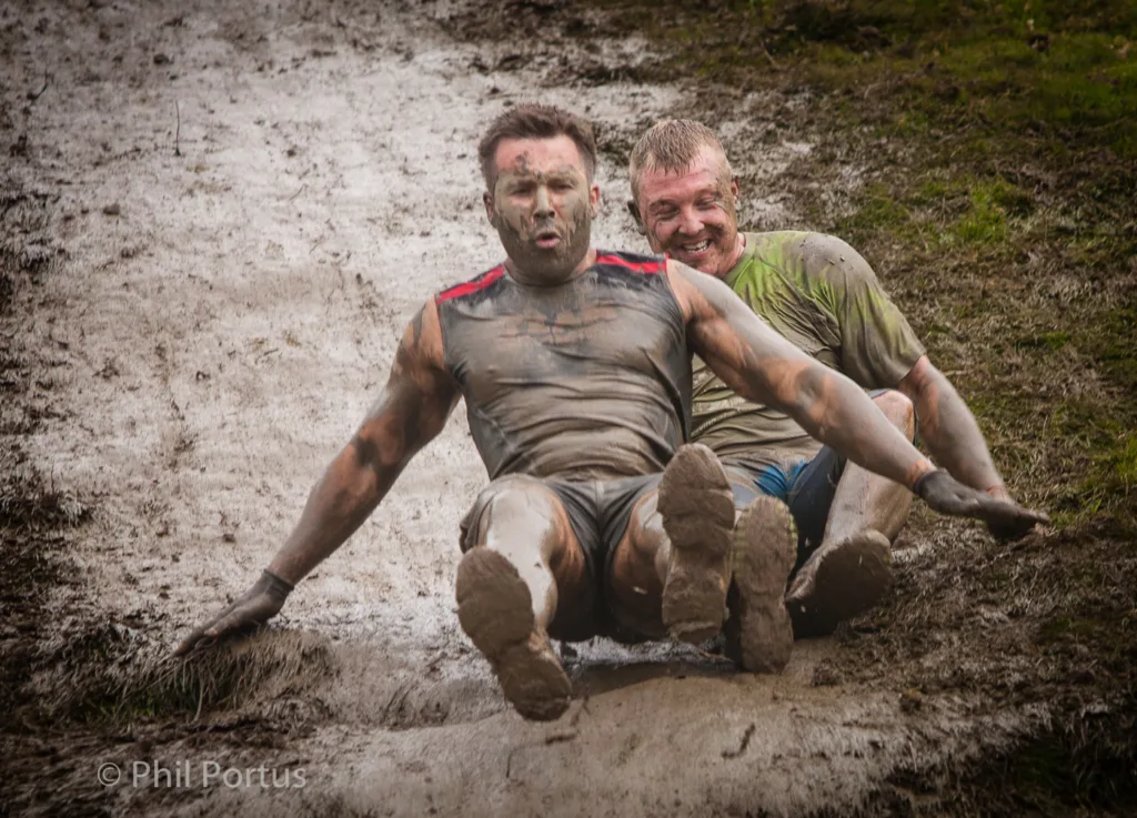 Two men covered in mud slide down a muddy slope, laughing and enjoying themselves.