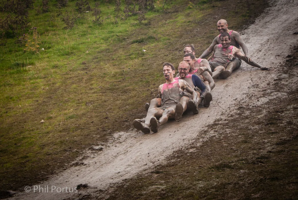 A group of people covered in mud slide down a grassy slope.