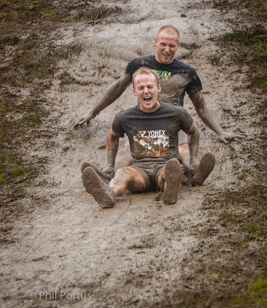 Two men covered in mud slide down a muddy slope, one pushing the other.