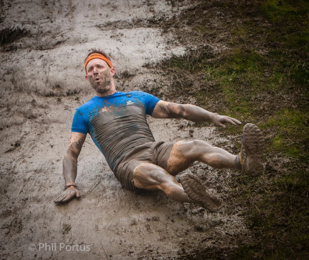 A person covered in mud lies on a muddy slope with their arms outstretched.