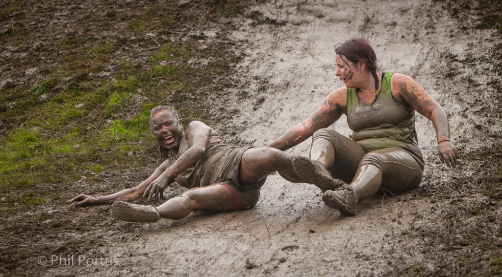 Two people covered in mud are playfully wrestling on a muddy slope.