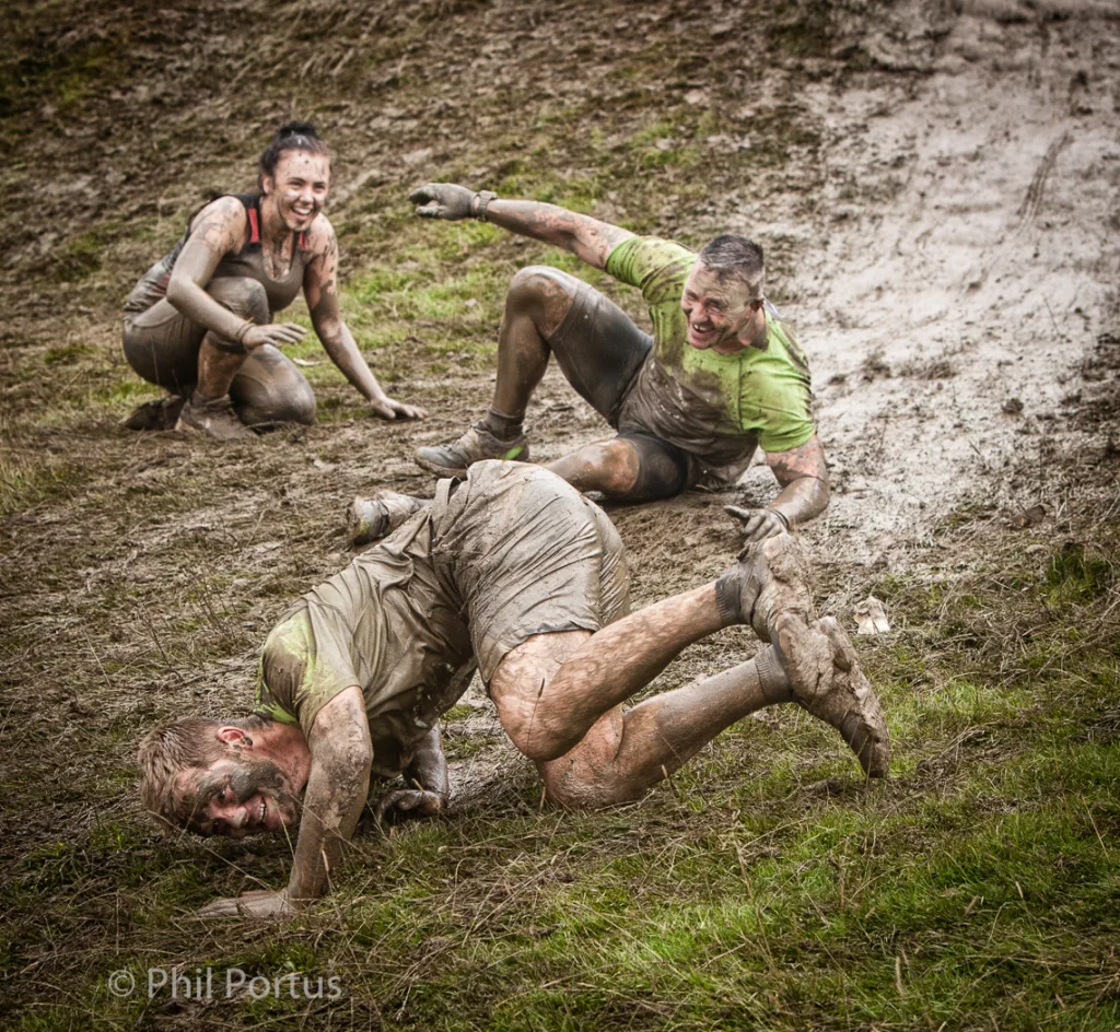 Three people covered in mud are laughing while one person falls forward on a grassy area.