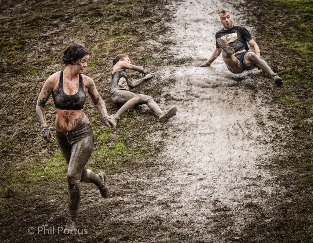 Three people covered in mud navigate a steep, muddy slope during an outdoor competition.