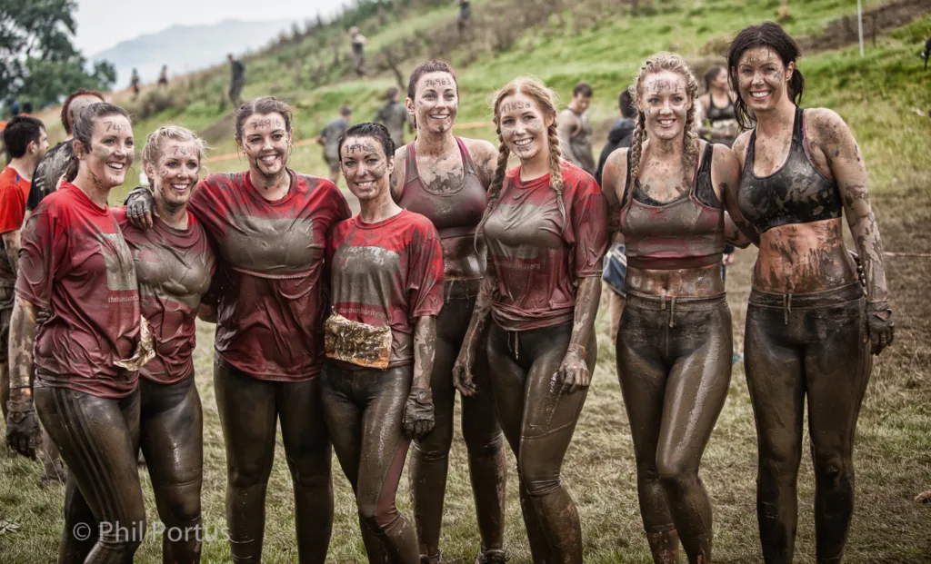 A group of seven people covered in mud smile for a photo on a grassy field.