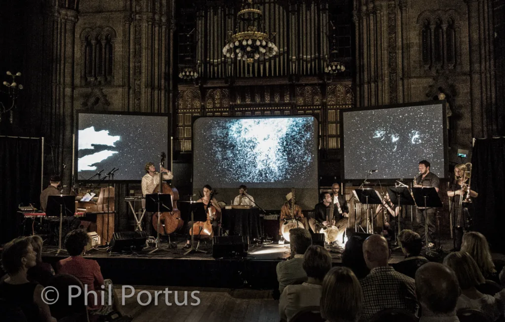 A musical ensemble performs on a stage with large screens displaying starry skies in a dimly lit building.