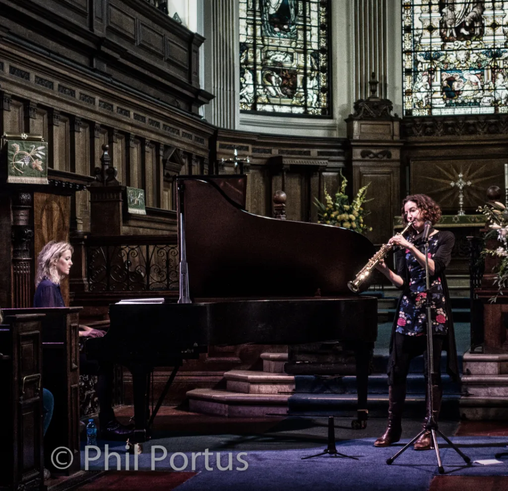 Two musicians perform on a stage with a grand piano and stained glass windows in the background.