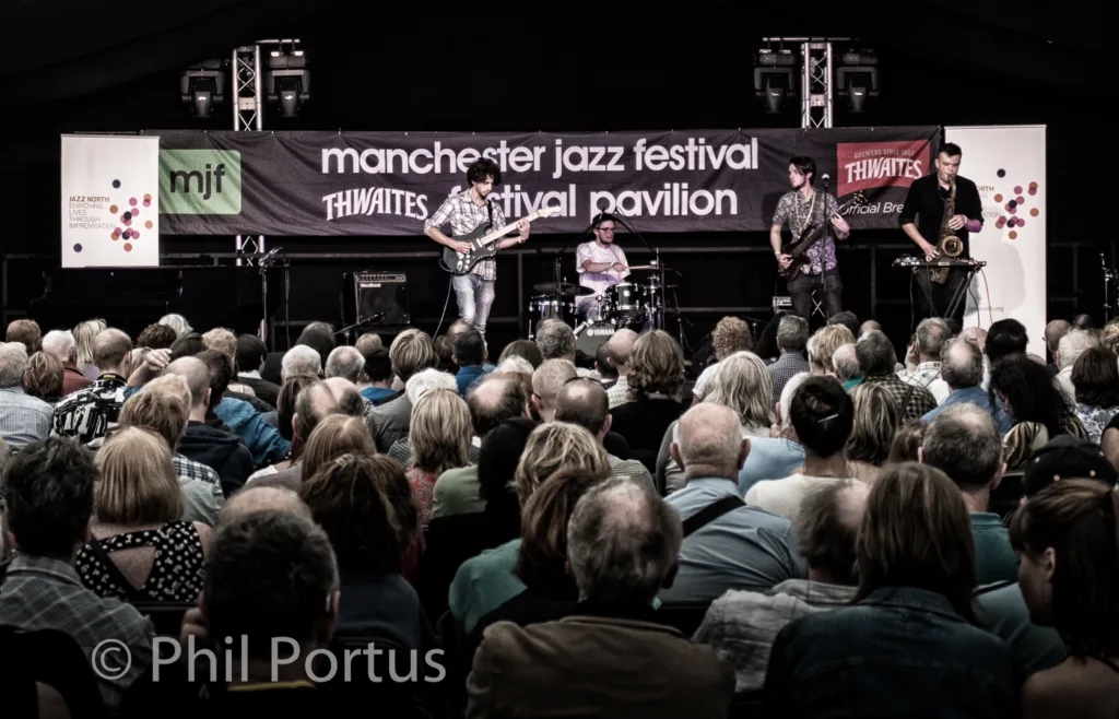 A band performs on a stage with a large crowd of people visible in the audience.