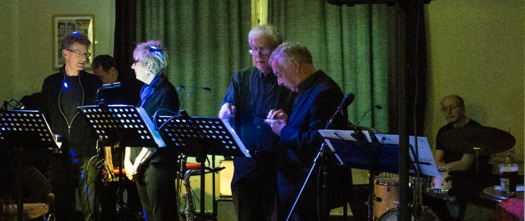 A jazz ensemble performs on a dimly lit stage with music stands and instruments visible.