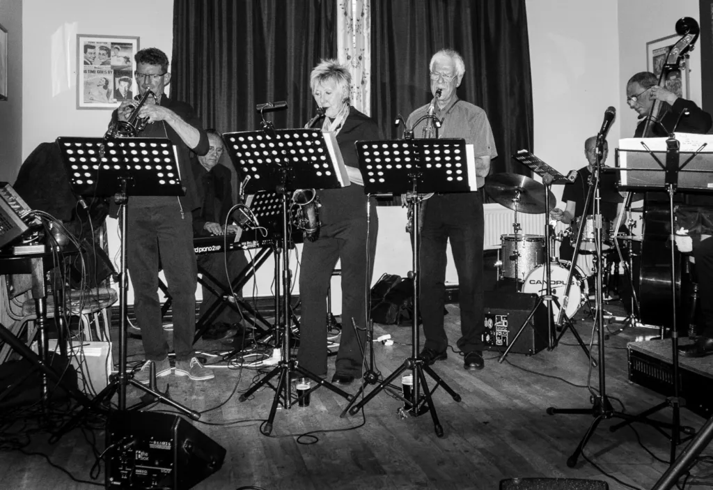 A black and white image shows a jazz band performing on a stage with music stands.