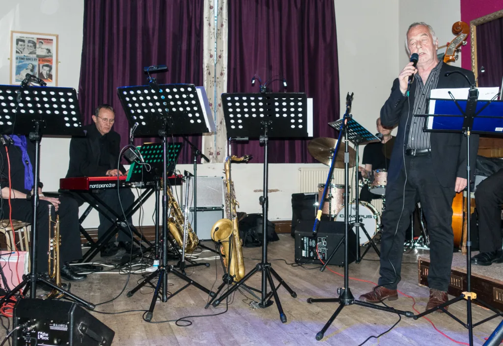 A band performs on a stage with music stands, instruments, and dark curtains in the background.