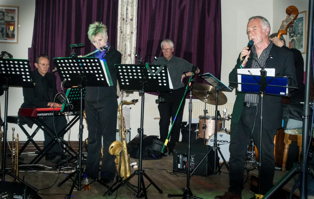 Four musicians perform on a stage with music stands and instruments visible.