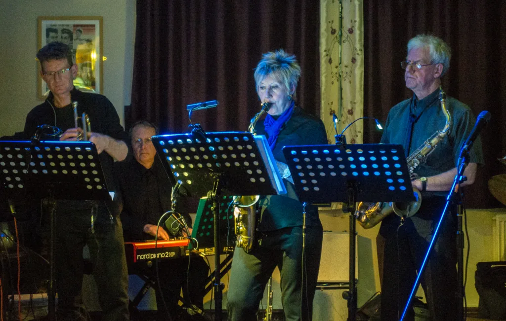 Four musicians perform on stage with music stands and instruments in a dimly lit room.