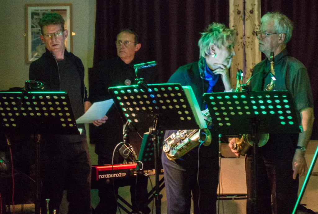 Four musicians stand on a dimly lit stage with music stands and instruments.