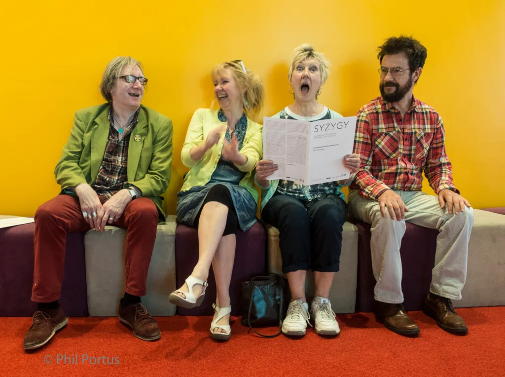 Four people sit on a purple bench against a yellow wall, reacting with surprise and amusement.