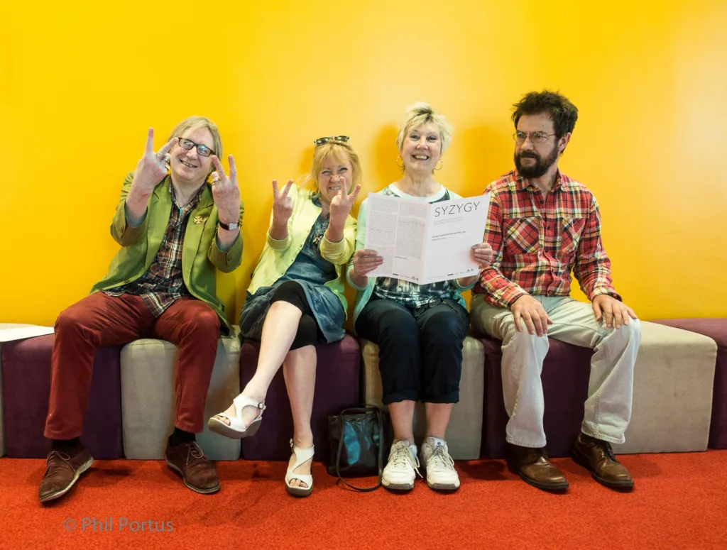Four people sit on a purple bench against a bright yellow wall, some making hand gestures.