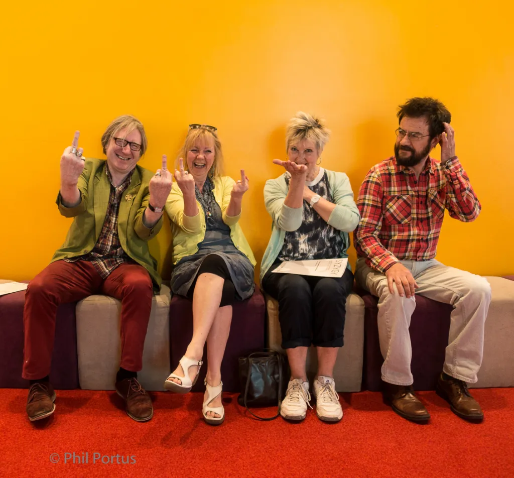 Four people sit on a grey couch against a bright orange wall, some making playful gestures.