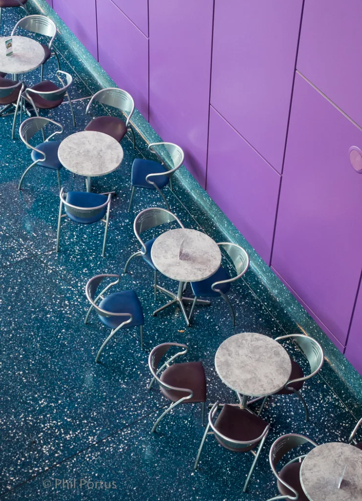 Round tables with chairs are arranged on a speckled blue floor next to a purple wall.