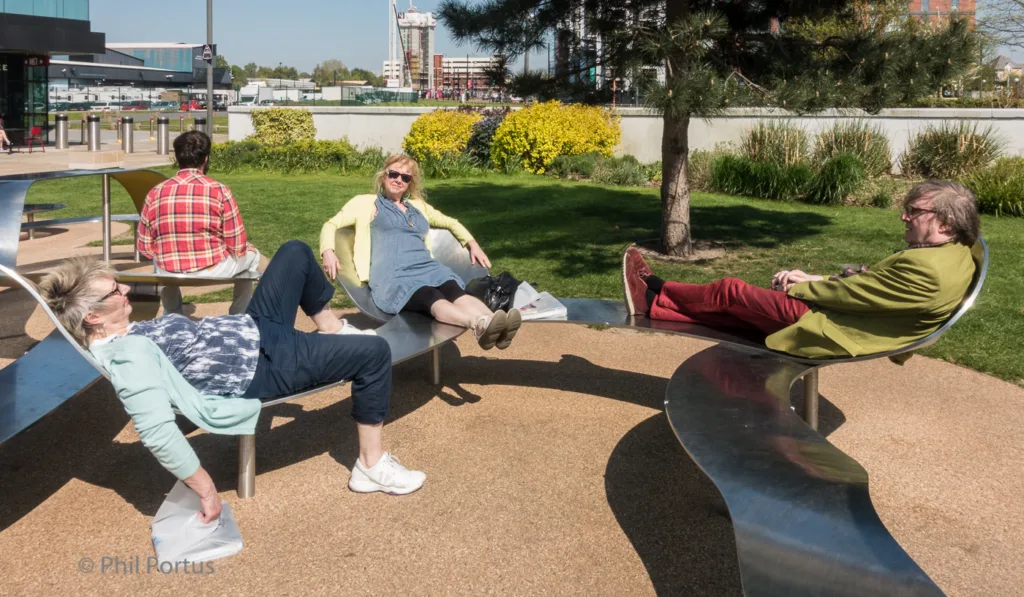 Four people relax on curved metal benches in a grassy outdoor area with modern buildings visible in the background.